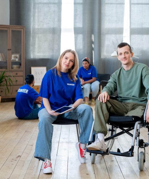 Young blond female sitting by disabled guy in wheelchair against group of volunteers